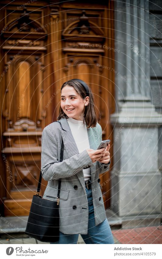 Positive Frau in Freizeitkleidung mit Smartphone auf einer Straße in der Stadt benutzend Großstadt Stehen laufen Browsen Surfen Mobile Telefon Lächeln Lachen