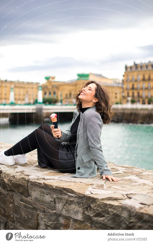 Zufriedene Frau genießt ein leckeres Eis am Kai Lächeln genießen Lachen Sitzen Zaun Hafengebiet Speiseeis Zapfen geschmackvoll bedeckt Großstadt Straße lässig