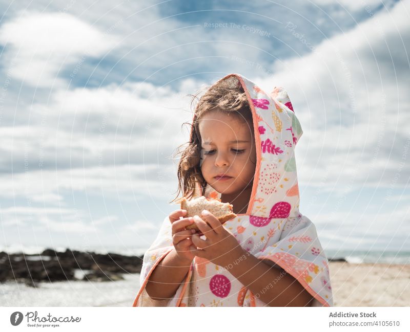 Kleines Mädchen isst Brot am Strand essen Sommer sonnig tagsüber wolkig Himmel Kind Kapuze Bekleidung Kleidungsstück Gebäck Brötchen Spielfigur frisch Kauen