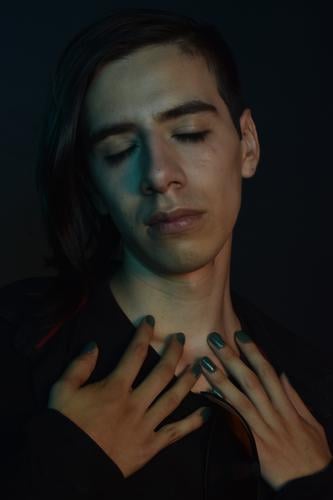 Junger gut aussehender Mann auf dunklem Hintergrund mit langen Haaren, geschlossenen Augen mit traurigem Ausdruck, Hände auf der Brust, grün lackierte Nägel