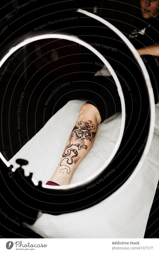 Anonyme Frau mit frischer Tätowierung bei Fototermin im Salon Tattoo Klient Fotoshooting Kosmetologie Haut Bein Stil Lampe Kultur professionell Design modern