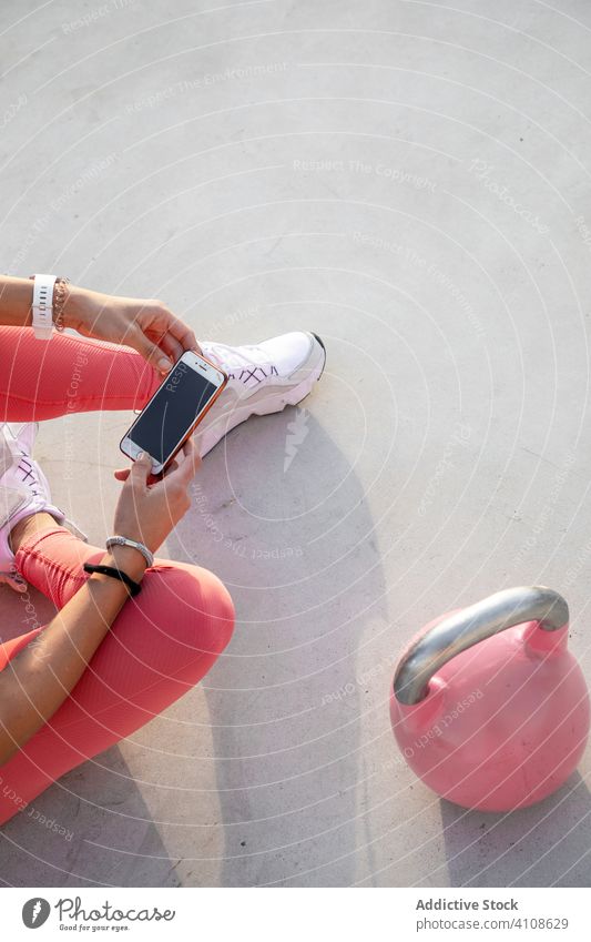 Anonyme Frau in rosa Leggings sitzt auf einem Dach und benutzt ein Smartphone Athlet Dachterrasse benutzend modern Kurzhantel ruhen Stock Browsen Pause