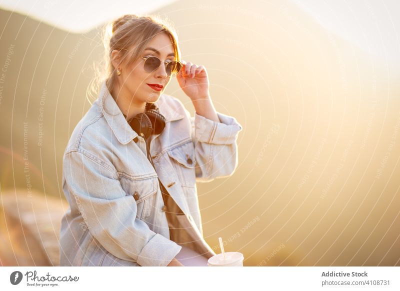 Wehmütige Frau benutzt Kopfhörer beim Entspannen ruhen sich[Akk] entspannen Natur sitzen Sonnenbrille Zaun lässig Getränk trinken benutzend Musik Stil Lifestyle