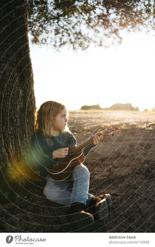 Kleines Mädchen sitzt unter einem Baum und spielt Ukulele Kind spielen Musik Gitarre Natur Sommer ernst wenig Instrument Klang Gesang Melodie sitzen Kofferraum