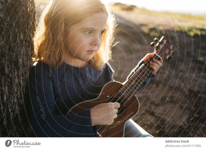 Kleines Mädchen sitzt unter einem Baum und spielt Ukulele Kind spielen Musik Gitarre Natur Sommer ernst wenig Instrument Klang Gesang Melodie sitzen Kofferraum