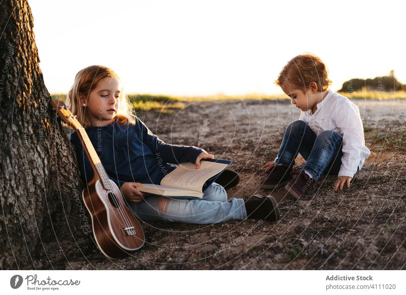 Kinder sitzen unter einem Baum und lesen ein Buch Natur Zusammensein Sommer Ukulele Gitarre ruhen ruhig Schwester Bruder Geschwisterkind Freund Landschaft