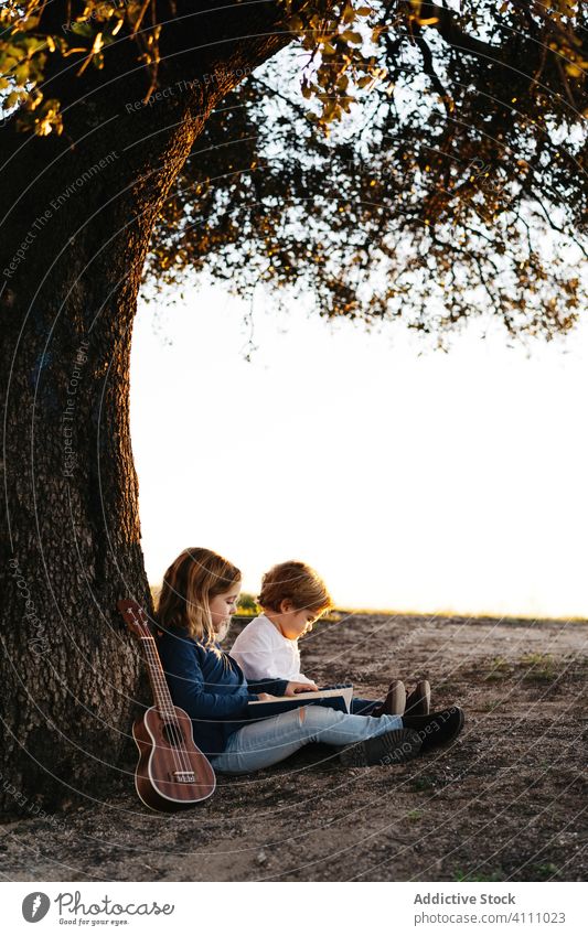 Kinder sitzen unter einem Baum und lesen ein Buch Natur Zusammensein Sommer Ukulele Gitarre ruhen ruhig Schwester Bruder Geschwisterkind Freund Landschaft