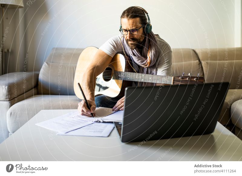 Zeitgenössischer Mann mit Gitarre schreibt Akkorde im Wohnzimmer Musiker Laptop Kopfhörer Liege schreiben Hinweis Songwriter Komponist Dichter männlich