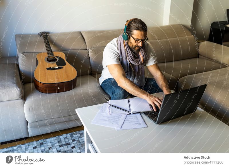 Konzentrierter Mann arbeitet mit Laptop im Wohnzimmer benutzend Kopfhörer Tippen Browsen zuhören Songwriter Brille männlich lässig zuschauen Anschluss online