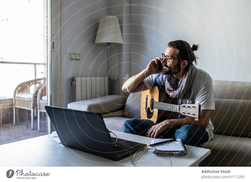 Junger männlicher Musiker, der im Wohnzimmer mit seinem Smartphone telefoniert reden Anruf Gitarre benutzend Browsen Laptop sitzen Liege Songwriter Komponist