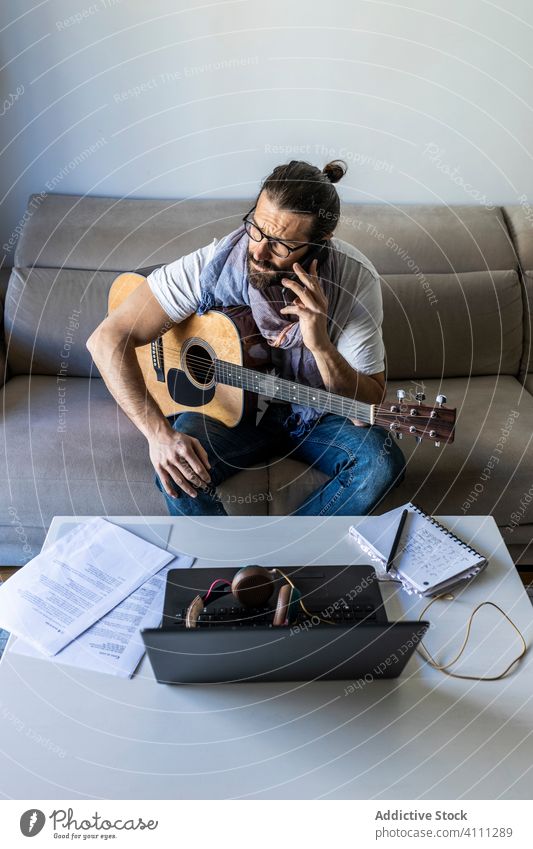 Junger männlicher Musiker, der im Wohnzimmer mit seinem Smartphone telefoniert reden Anruf Gitarre benutzend Browsen Laptop sitzen Liege Songwriter Komponist