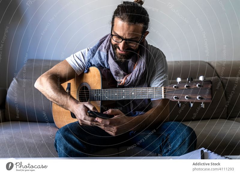 Fröhlicher Mann mit Gitarre, der auf dem Sofa sein Smartphone überprüft sitzen Liege benutzend Browsen Lächeln männlich lässig Musiker Mobile Telefon Vollbart