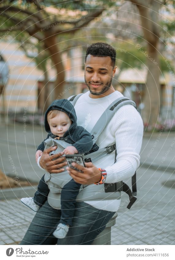 Glücklicher schwarzer Mann mit Baby in der Trage und Telefon auf der Straße Vater Smartphone benutzend Träger Texten urban modern Papa Kind Säugling Sohn