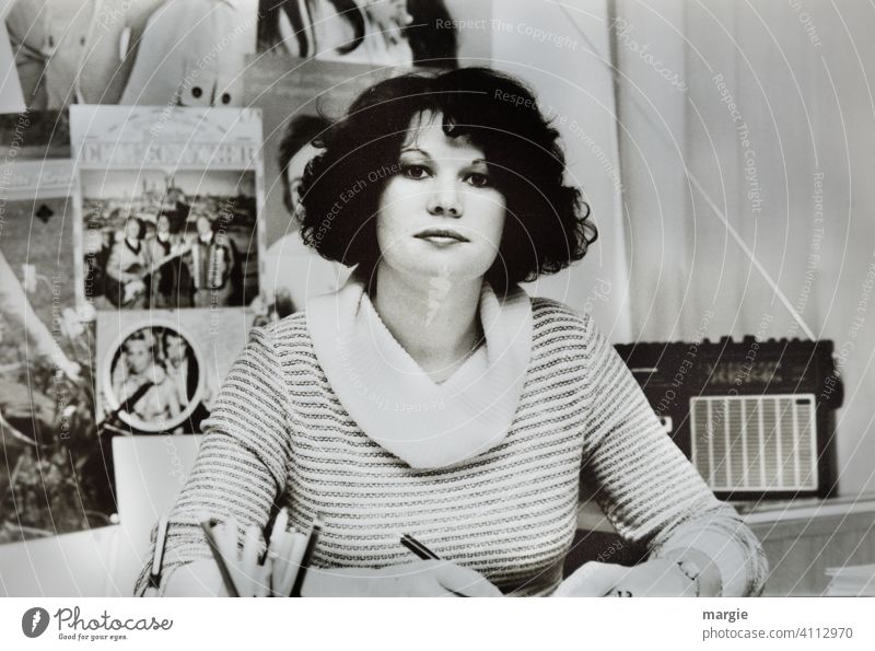 Eine junge Frau sitzt im Büro an ihrem Schreibtisch und schaut in die Kamera. Hinter ihr steht ein altes Radio. Künstlerfotos hängen an der Wand. Junge Frau