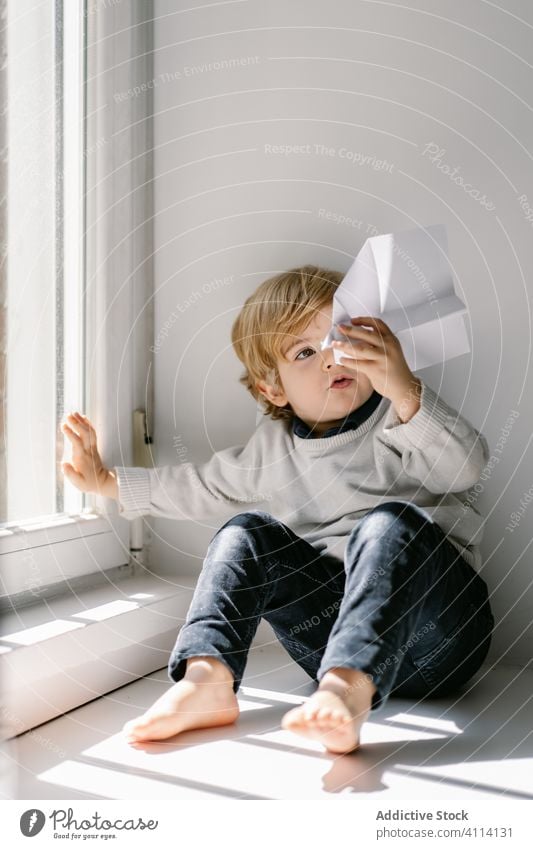 Neugieriger kleiner Junge spielt mit Papierflugzeug auf weißer Fensterbank zu Hause spielen Papierflieger Fensterbrett träumen heimwärts neugierig unschuldig