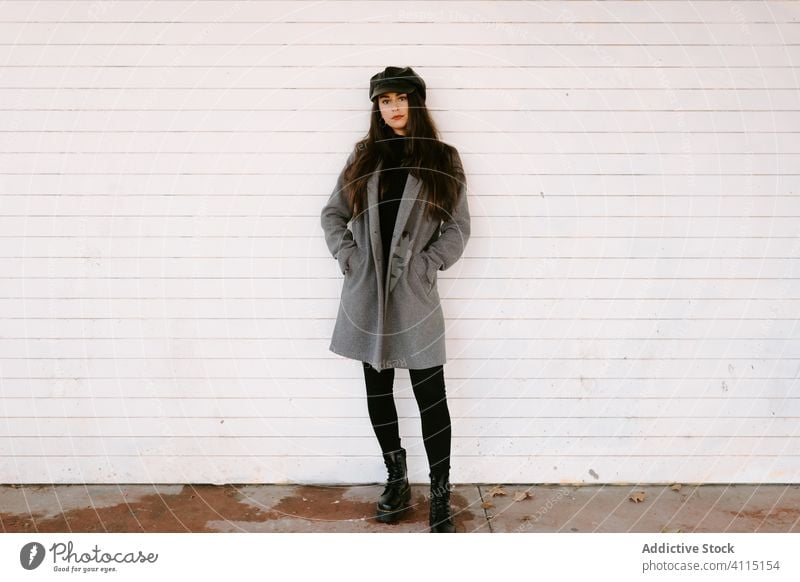 Selbstbewusste junge Frau an der weißen Wand Stil Straße selbstbewusst Verschlussdeckel Gebäude Mantel Outfit Model ernst urban anhaben Accessoire Hut brünett