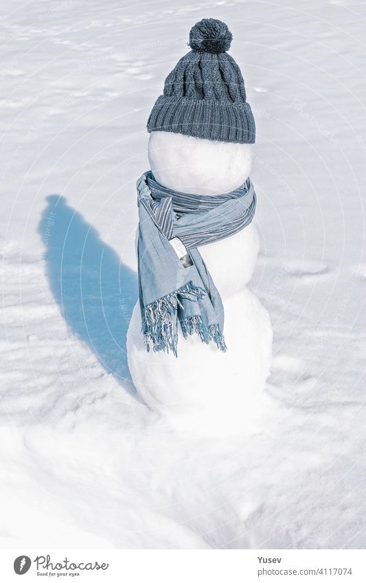 Ein Schneemann mit blauer Mütze und Schal steht im weißen Feld. Ein Eismann auf einem hellen, auffälligen Hintergrund. Sonniger Tag, eiskalt, Nahaufnahme. Hut