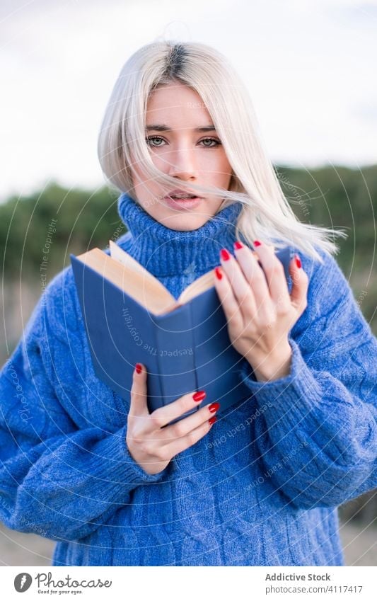 Junge Frau mit Buch schaut in die Kamera blau Pullover lesen jung lässig ernst Natur blond selbstbewusst modern Bildung Hobby Schüler ruhen klug lernen Wissen