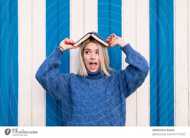 Erstaunte junge Frau mit Buch auf dem Kopf Spaß erstaunt blau Wand lässig Pullover gestrickt wow Erstaunen Farbe hell farbenfroh Stil trendy modern aufgeregt