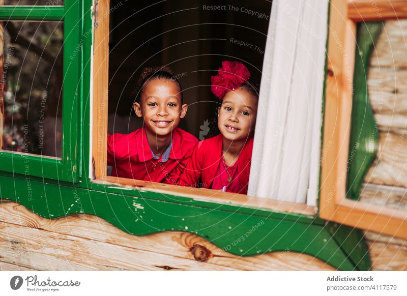 Fröhliche ethnische Kinder schauen aus dem Fenster Haus Glück Geschwisterkind Zusammensein heiter hölzern ländlich Lächeln heimwärts Lifestyle Landschaft Freude