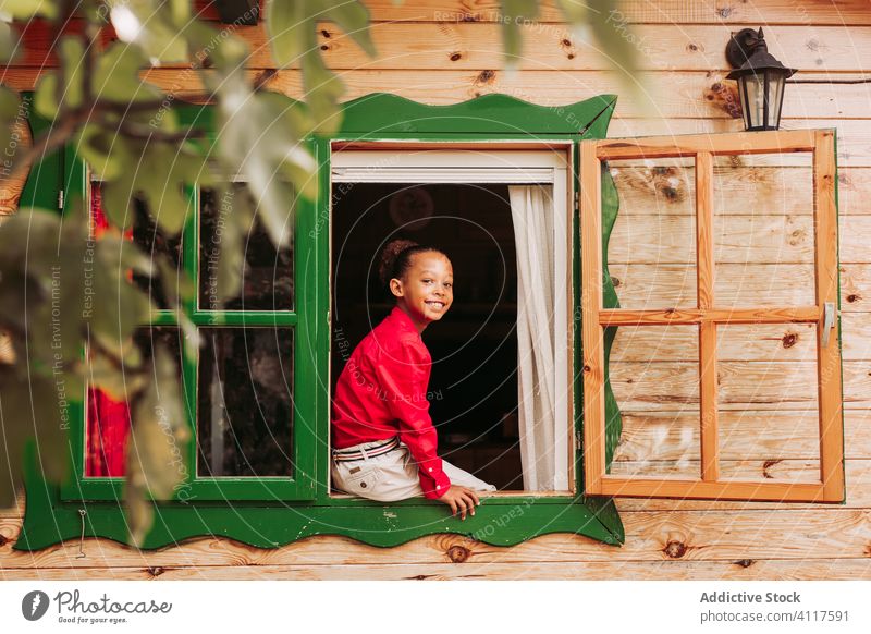 Glückliches ethnisches Kind schaut aus dem Fenster Haus heiter hölzern ländlich Lächeln heimwärts Lifestyle Landschaft Freude wenig lässig Kindheit schwarz