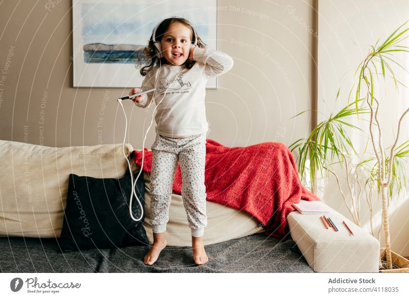 Niedliches kleines Mädchen springt auf der Couch und hört Musik mit Kopfhörern niedlich wenig posierend heimwärts im Innenbereich Fenster Kind Porträt Kindheit