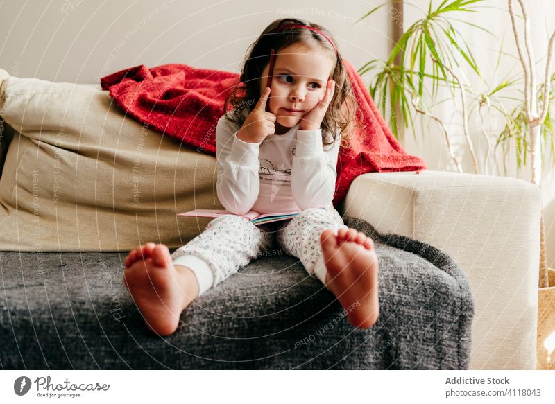Nettes kleines Mädchen sitzt auf der Couch mit einem Notizbuch auf der Suche nach Inspiration niedlich wenig posierend heimwärts im Innenbereich Fenster Kind