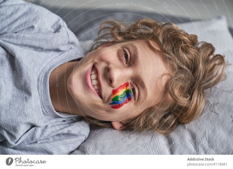 Glücklicher Junge mit Regenbogen im Gesicht schaut in die Kamera heimwärts Quarantäne auf die Kamera zeigen Konzept Symbol Lächeln Lügen Stock Decke Kopfkissen
