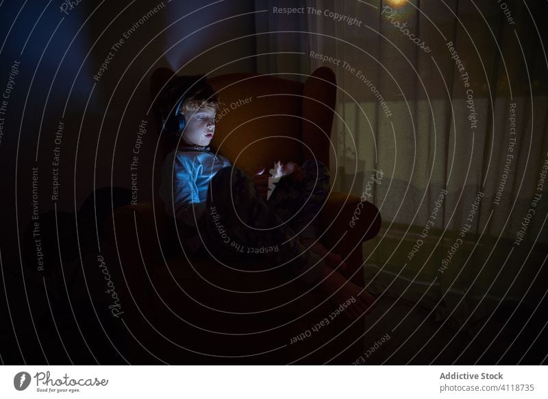 Kind sitzt in einem Sessel in einer Zimmerecke und schaut nachts mit Kopfhörern auf ein Tablet Junge Schutzhelm Musik zuhören Spiel spielen Gerät Tablette