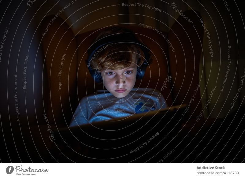 Kind sitzt mit Kopfhörern auf einem Sofa und schaut nachts auf das Tablet Junge interagieren Verkleidung Gerät elektronisch Vorstellungskraft Multimedia