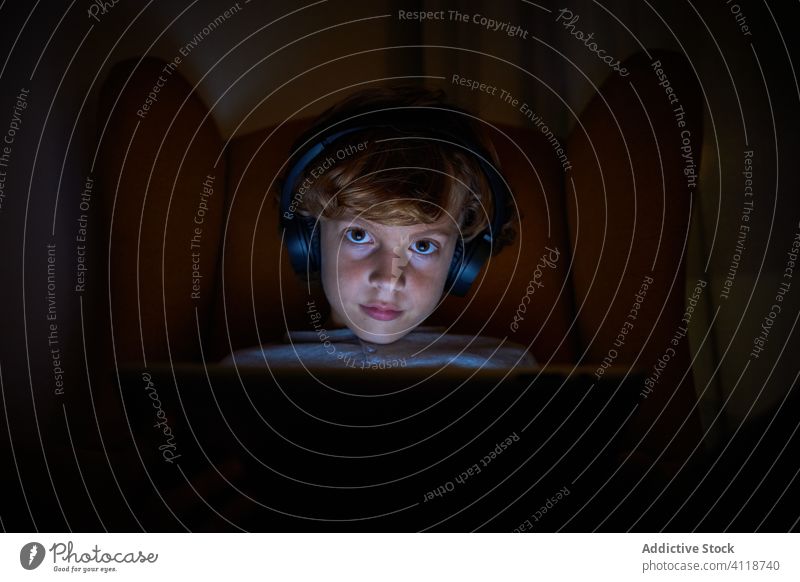 Kind sitzt mit Kopfhörern auf einem Sofa mit Tablet-Computer und schaut nachts frontal Junge Abgelenkt Vorstellungskraft Kino Multimedia Mitteilung zuhören