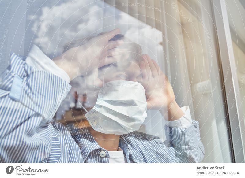 Junge hinter Fenster während der Quarantäne Mundschutz medizinisch heimwärts Stirnrunzeln Pandemie Coronavirus Bund 19 Kind Kindheit traurig unglücklich Hygiene