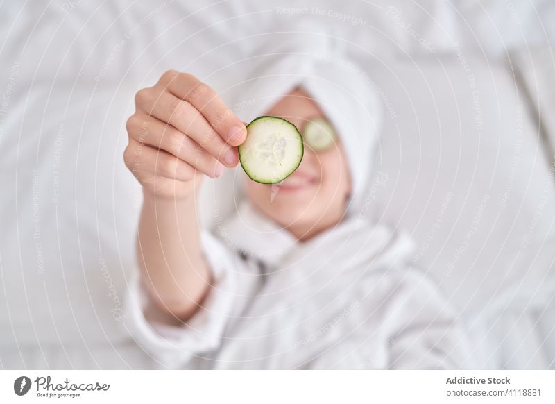 Glückliches Kind genießt die Schönheitsbehandlung Salatgurke Mundschutz Hautpflege Spaß positiv genießen zeigen Scheibe Gesichtsbehandlung Witz Humor Bett