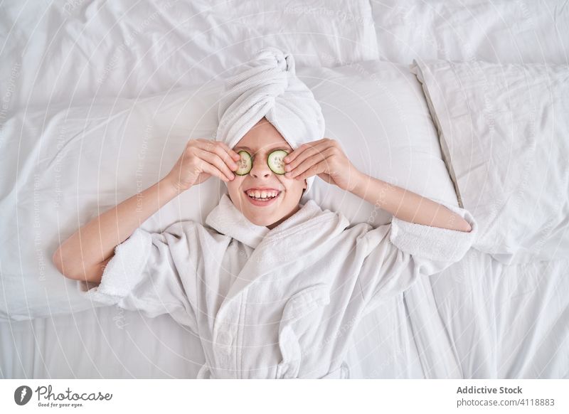 Fröhliches Kind mit Gurkenscheiben auf ja auf dem Bett liegend Salatgurke Mundschutz Schönheit Hautpflege Spaß positiv Lachen genießen Gesichtsbehandlung Witz