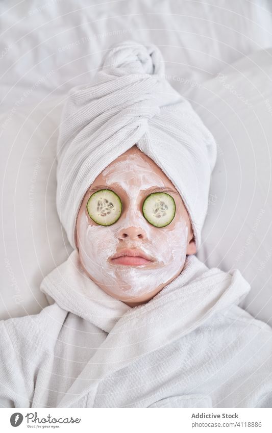 Verspieltes Kind mit Gesichtsmaske auf dem Bett liegend Mundschutz Gesichtsbehandlung Spaß haben Salatgurke Schönheit Hautpflege Grimasse spielerisch