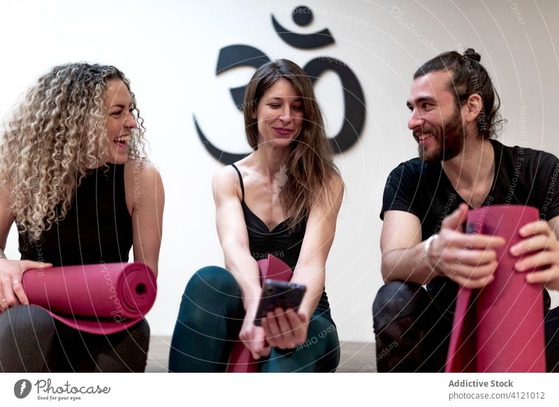 Fröhliche Freunde nutzen ihr Smartphone vor dem Yoga-Training benutzend Atelier Lächeln teilen vorbereiten Mann Frauen sitzen Zusammensein Fitnessstudio heiter