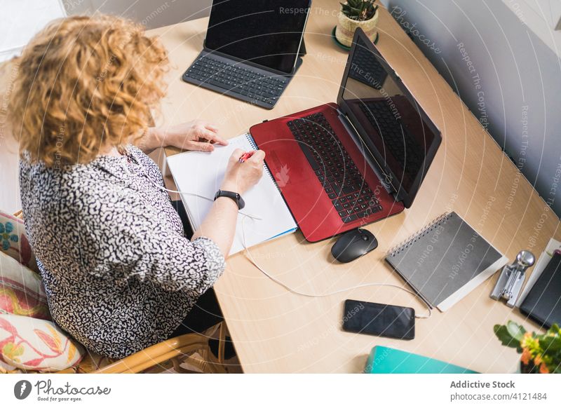 Anonyme Frau macht sich Notizen in einem Notizbuch, während sie an einem Laptop arbeitet Notebook Smartphone Arbeitsplatz heimwärts freiberuflich abgelegen
