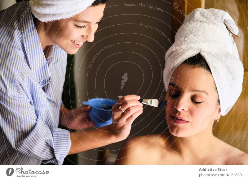 Glückliche Frau, die eine Tonmaske auf das Gesicht einer Freundin aufträgt Frauen Kälte heimwärts Schönheit Verfahren Hautpflege zufrieden bewerben