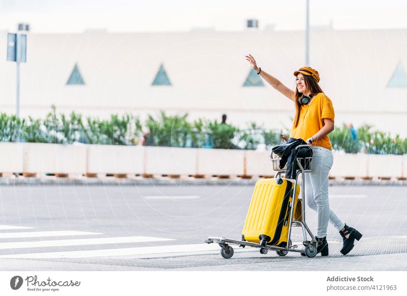 Glückliche Frau mit Gepäckwagen, die ein Taxi in der Nähe des Flughafens nimmt Karre Hagel Straße fangen Stil Gebäude Kopfhörer Verkehr Passagier