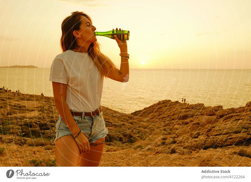 Lächelnde junge Frau mit Bierflasche bei Sonnenuntergang am Meer Reisender Meeresufer Erfrischung Sommer Kälte Freizeit Glück Ibiza Spanien Sonnenbrille Flasche