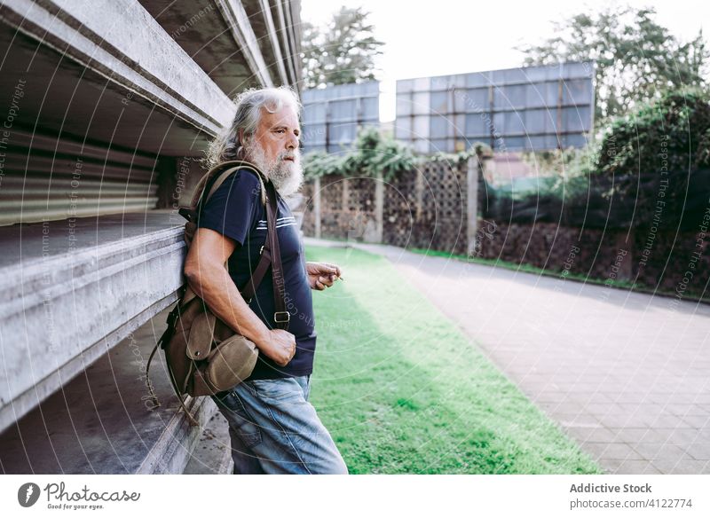Älterer reisender Mann, der auf der Straße raucht Reisender gealtert Großstadt männlich Tourist älter lange Haare graues Haar Senior lässig anhaben Rucksack