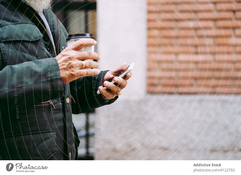 Älterer Mann mit Kaffee zum Mitnehmen und Smartphone beim Spaziergang gealtert schlendern Imbissbude Browsen Straße benutzend männlich Heißgetränk Senior älter