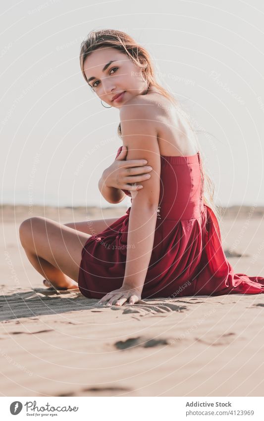 Attraktive junge Frau mit langen blonden Haaren trägt stilvolle rote Kleid sitzt auf Küste und schaut in die Kamera Strand sinnlich Sand Stil Sommer allein