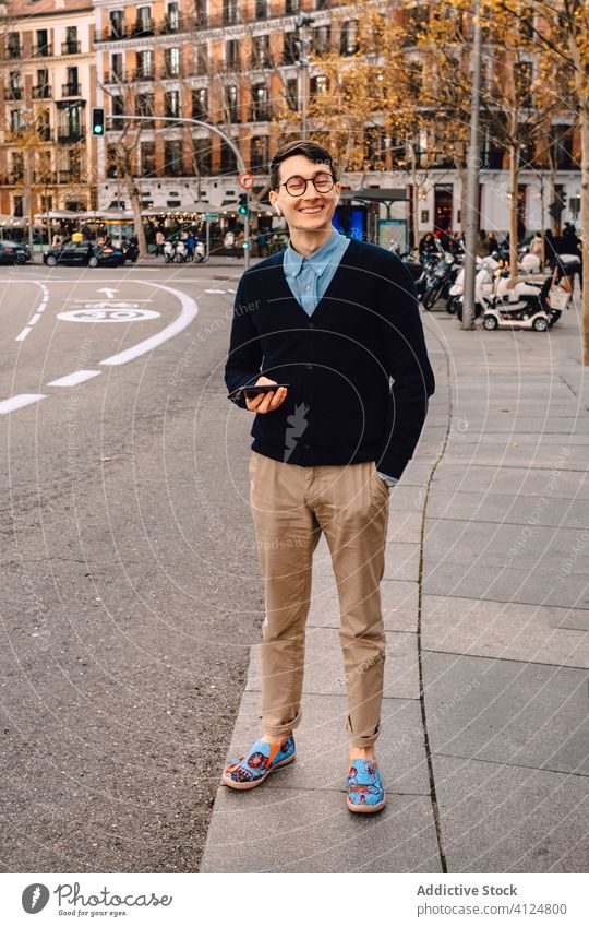 Stilvoller junger Mann mit Smartphone auf dem Gehweg stehend trendy Straße Mode selbstbewusst Musik Bürgersteig Brille Kopfhörer Großstadt Outfit Gerät