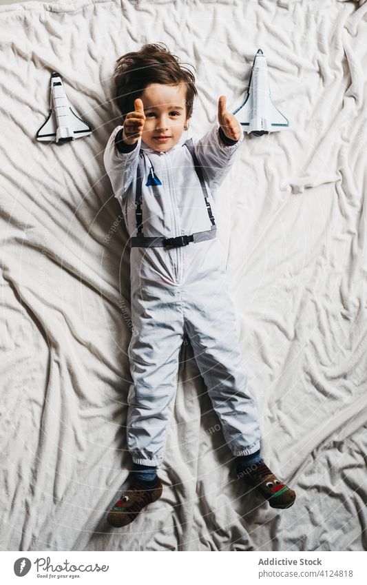 Junge im Astronautenkostüm auf dem Bett liegend Tracht Raumanzug Lügen Spielzeug Raumschiff Spaß haben Lächeln Kind Kosmonaut Schlafzimmer heiter Wochenende