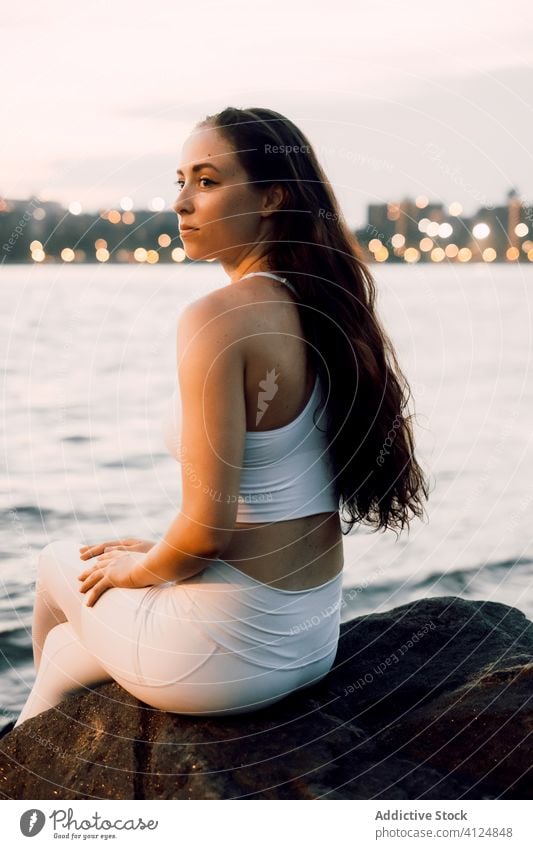 Ruhige Frau in Lotus-Pose am felsigen Ufer eines Flusses Stadtbild Küste Stein Hafengebiet üben Abend Windstille Konzentration Fokus ruhig Gelassenheit