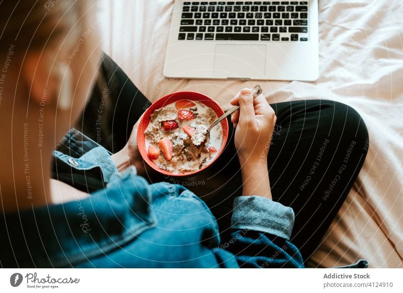 Frau isst Müsli mit Erdbeeren und benutzt Laptop im Bett heimwärts essen benutzend Kopfhörer zuschauen soziale Netzwerke Filmmaterial Video zuhören Surfen