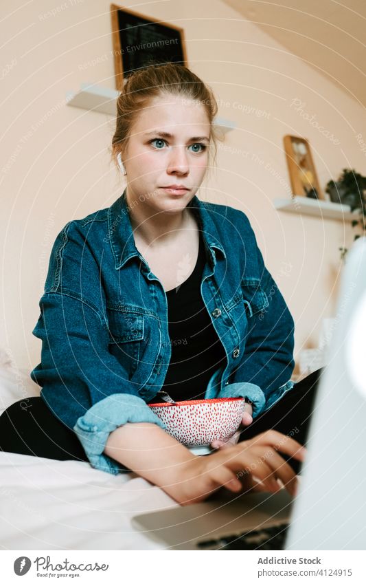 Schockierte junge Frau mit Ohrstöpseln und Laptop beim Mittagessen in einem hellen, modernen Schlafzimmer zu Hause heimwärts benutzend Kopfhörer Bett lesen