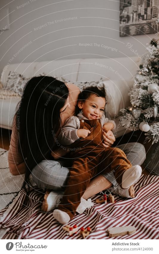 Fröhliche, liebevolle Mutter umarmt kleinen Sohn, der auf dem Boden neben dem Weihnachtsbaum sitzt kuscheln Weihnachten heimwärts spielerisch gemütlich Feiertag