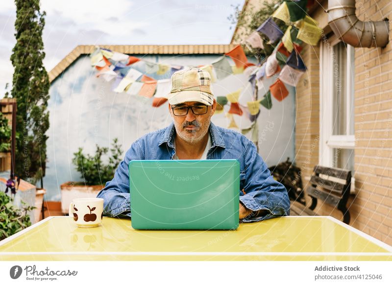 Nachdenklicher älterer Mann in Gläsern mit heißem Getränk und Laptop auf der Terrasse eines Landhauses trinken besinnlich lesen zuschauen verwenden Becher